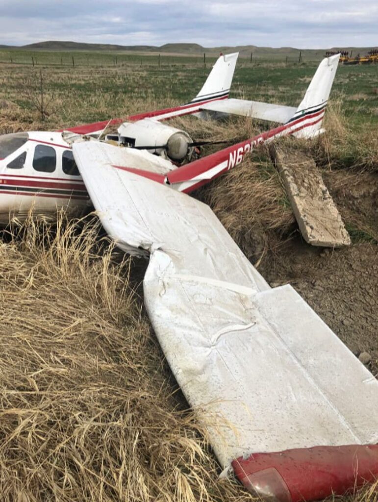 View of the damaged left wing and tail of Cessna P337H N62PC resting in an irrigation ditch, showing crumpled wing and broken tail structure.